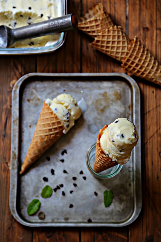 mint chocolate chip ice cream cone laying on baking sheet with chocolate chips scattered. Ice Cream cone standing in glass jar on baking sheet. Container of ice cream and cones in background.