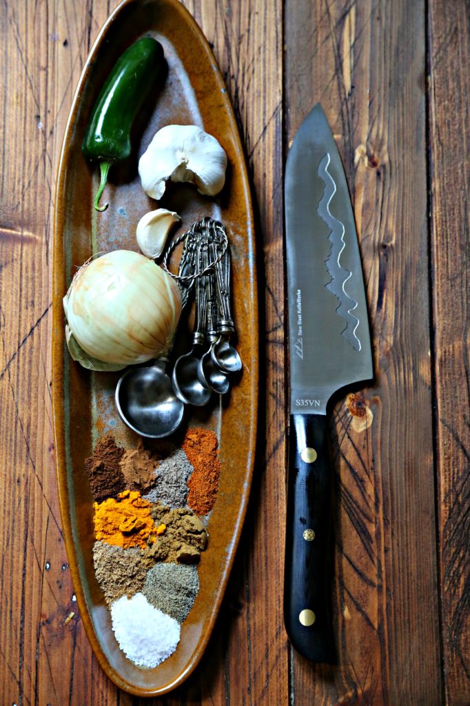 Brown oblong tray with ingredients (jalapeno, onion, garlic, spices and measuring spoons). Knife to right. 