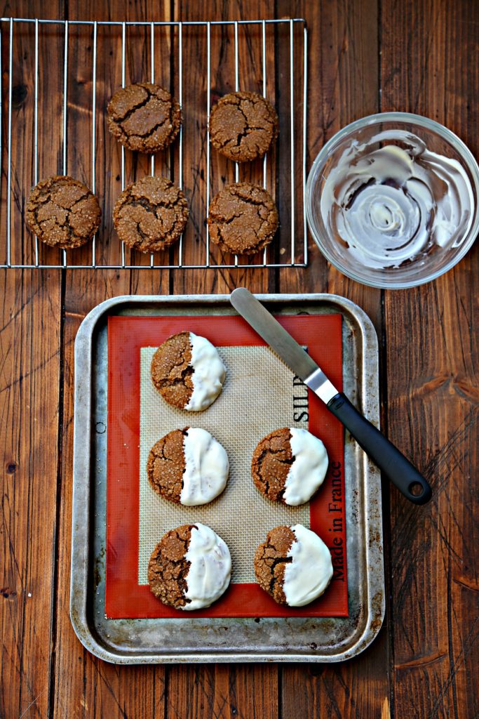 White Chocolate Dipped Chewy Gingersnap Cookies on baking sheet. Cooling rack with cookies and glass bowl with chocolate in back.