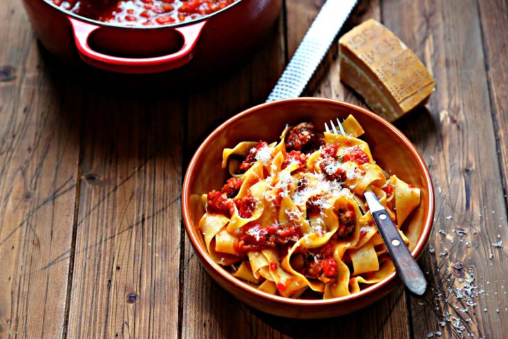 brown bowl with pasta and meat sauce. Red pot with sauce and wedge of cheese behind.