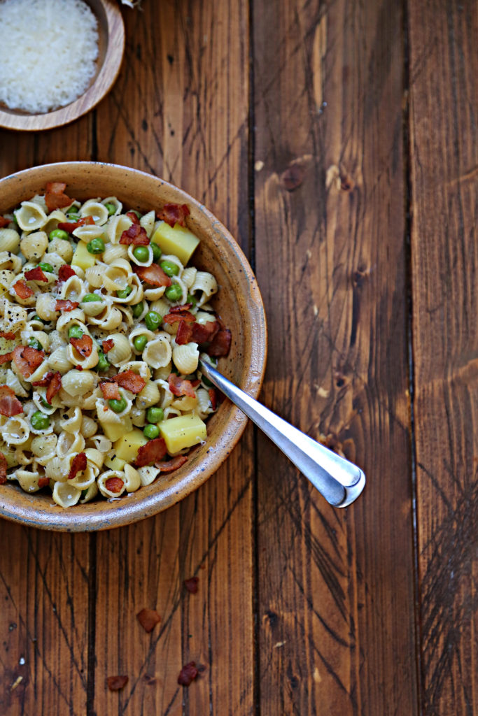 partial view of brown bowl with shells pasta, peas and bacon. Serving spoon handle visible.