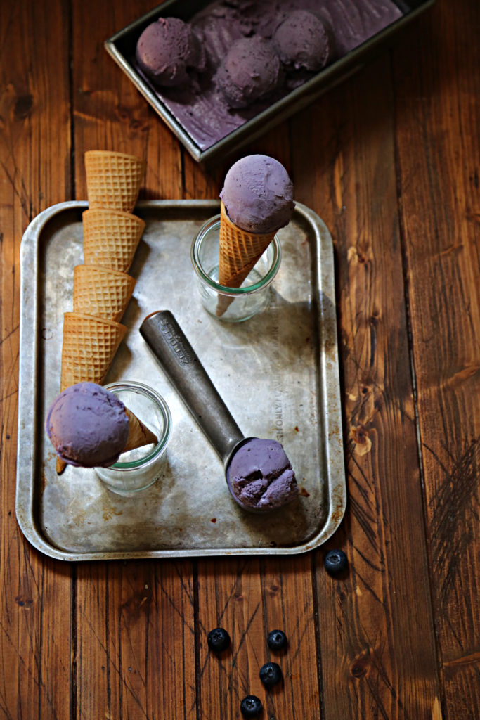 baking sheet with stack of sugar cones, 2 small glass jars with ice cream cones. Ice cream scooper with scoop of blueberry ice cream.