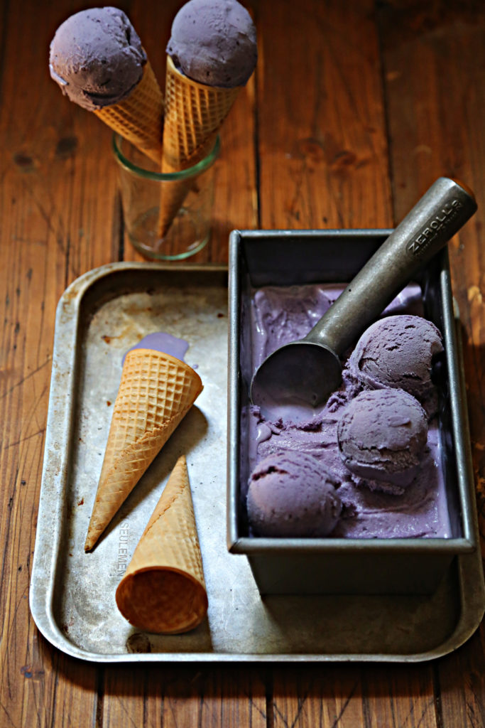 baking sheet with sugar cones and loaf pan of blueberry ice cream. 2 scooped ice cream cones in glass behind.