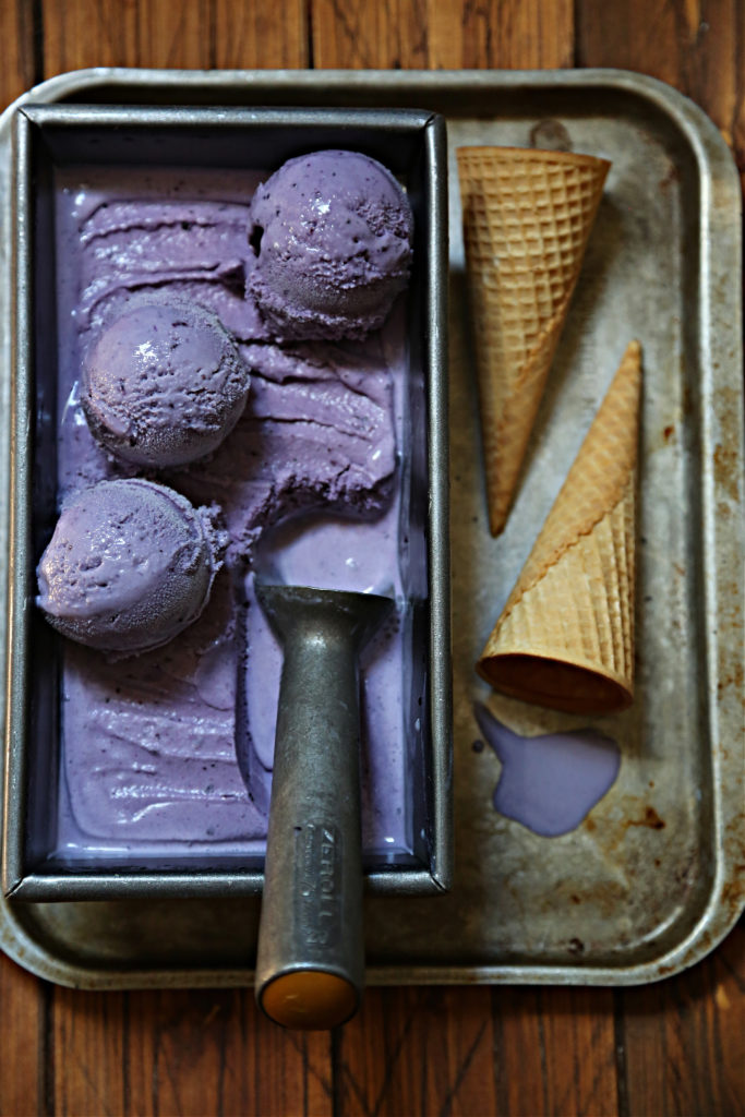 loaf pan of blueberry ice cream, scoop on top. 2 sugar cones to right of pan on baking sheet.