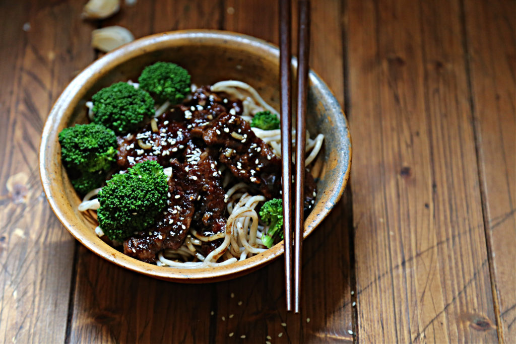 brown bowl with noodles broccoli and beef teriyaki. Chopsticks resting on bowl. Garlic cloves behind bowl.