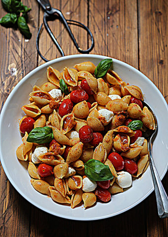 white bowl with pasta shells, tomatoes, basil and mozzarella balls. Serving spoon in bowl. Scissors and basil surrounding.