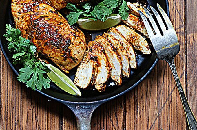 2 cooked chicken breasts, 1 sliced with scattered cilantro and lime wedges in black skillet. Fork resting on side.
