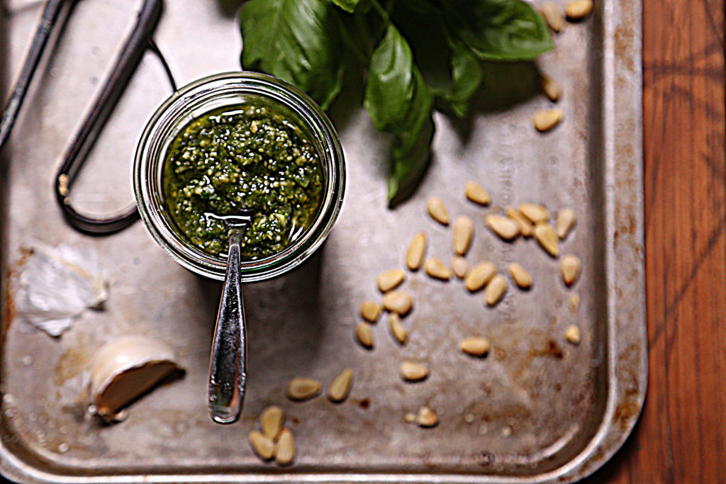 glass jar of pesto on baking sheet with scattered pine nuts, basil leaves and scissors.