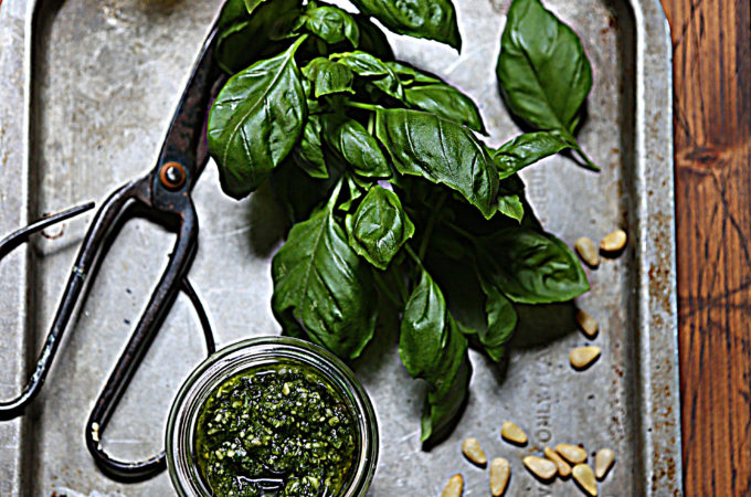 pesto in jar with spoon on baking sheet with basil, pine nuts, scissors and garlic cloves. Glass jar of olive oil and cheese grater in background.