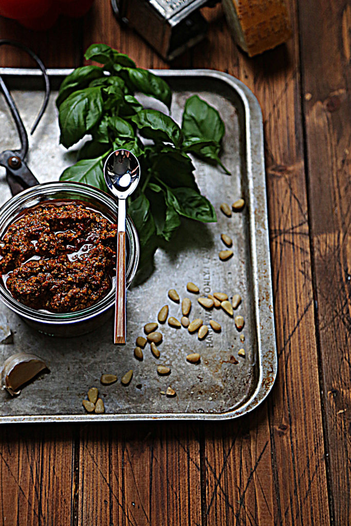 baking sheet with basil, pine nuts, scissors, glass jar of red pesto and spoon.