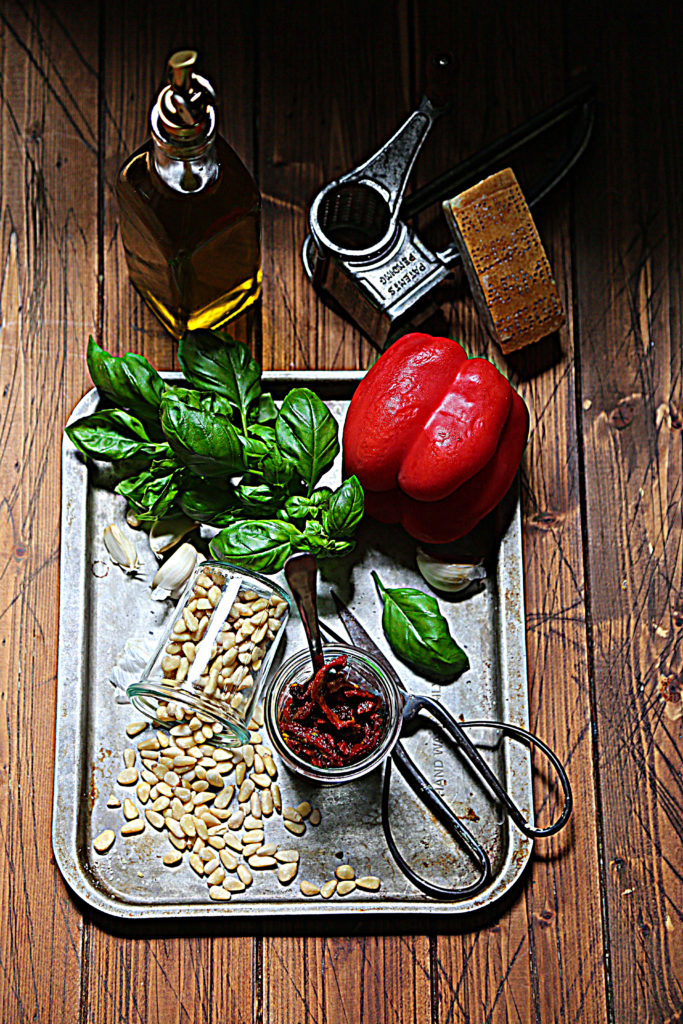baking sheet with basil, pine nuts, bell pepper, scissors and jar of sun dried tomatoes with spoon. Jar of olive oil and cheese grater behind baking sheet.