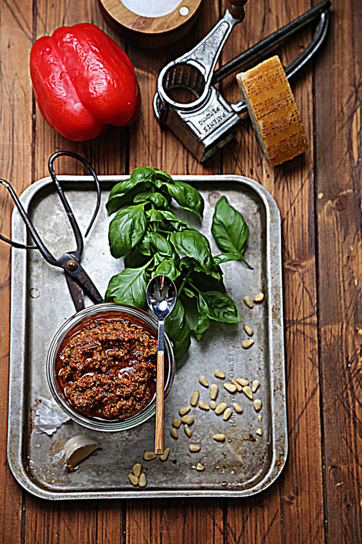baking sheet with jar of red pesto and spoon. Basil, pine nuts and scissors on baking sheet. Red bell pepper and cheese grater in background.