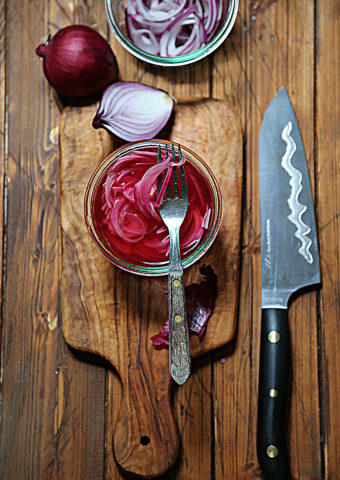 glass jar of pickled onions with fork. Knife to side. Whole onion and half onion in background.