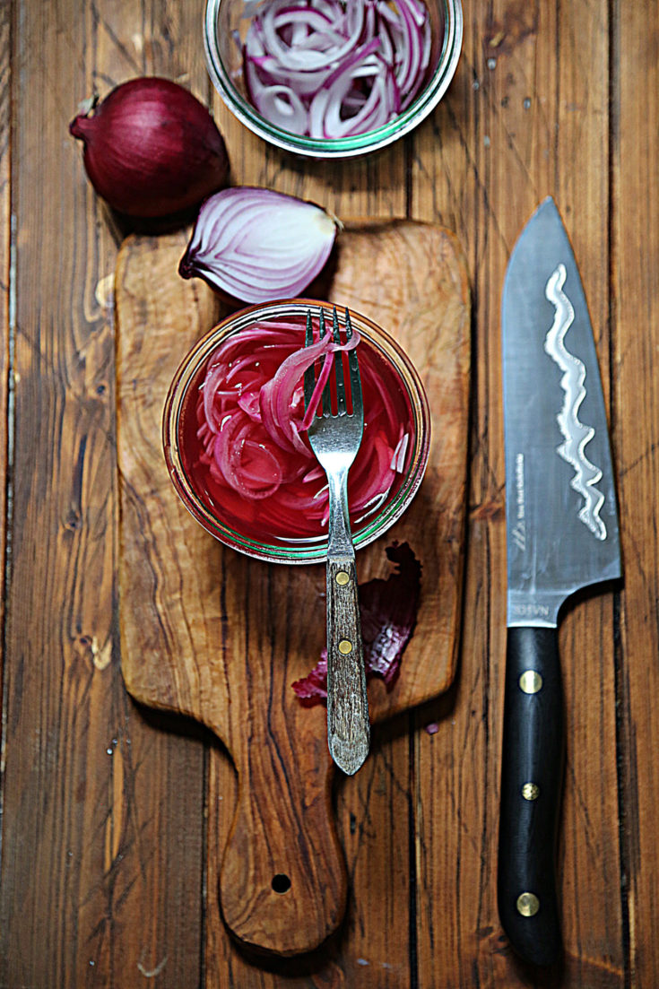 glass jar of pickled onions with fork. Knife to side. Whole onion and half onion in background.