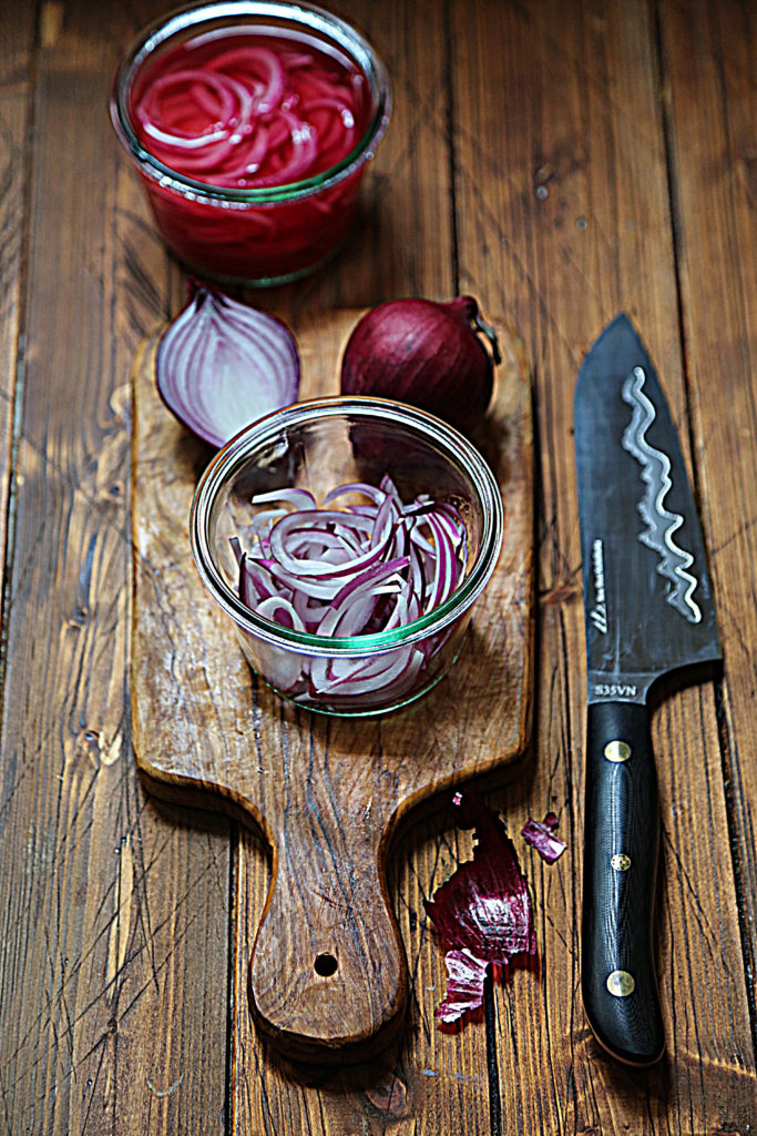 glass jar of sliced red onions. Whole onion, half onion and jar of pickled onions in background. Knife to side. 