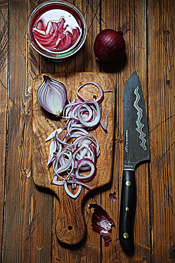 cutting board with half of red onions and slices of onions. Knife to right. Small jar of pickled onions and whole onion above board. 
