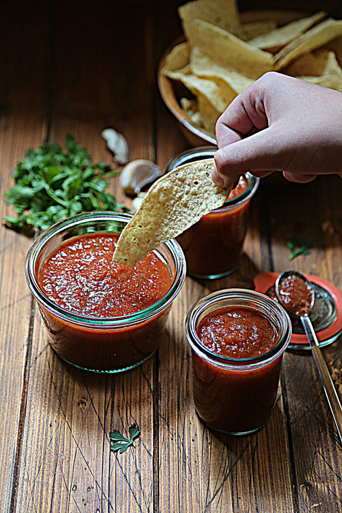 hand dipping chip into jar of salsa. cilantro, garlic cloves and 2 other jars of salsa surrounding. 