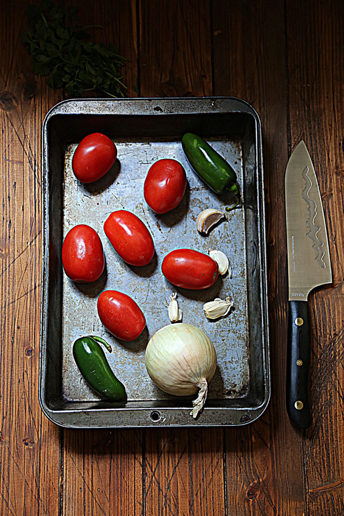 baking sheet with tomatoes, jalapenos, garlic cloves and onion. Knife to side of pan.