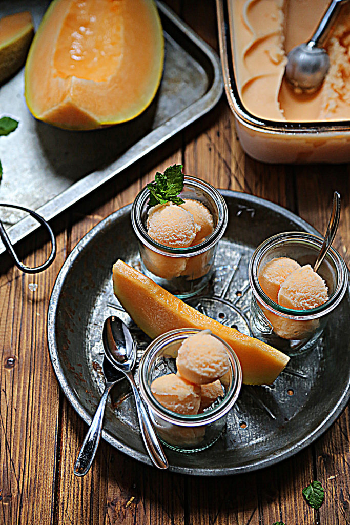 tray of small glass jars of cantaloupe sorbet. Sliced cantaloupe on baking sheet in background. 