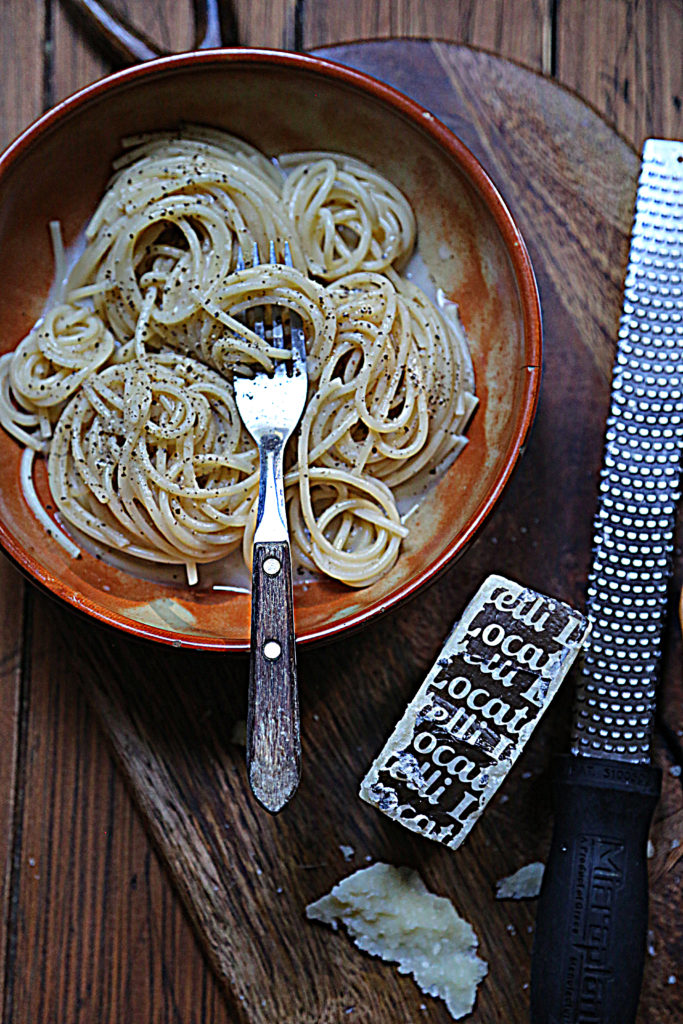 brown bowl of cacio e pepe pasta with fork. Microplane and cheese wedge to side. 