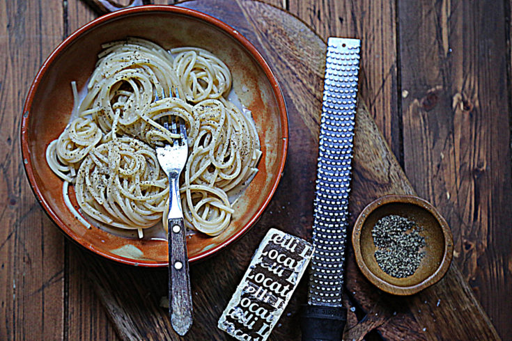 bowl of cacio e pepe pasta with fork. Bowl of pepper, microplane and wedge of Pecorino cheese to side.