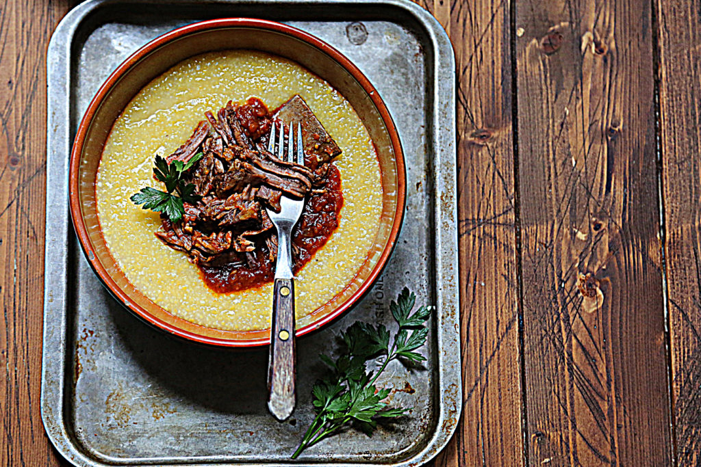 brown bowl with creamy polenta and shredded short ribs sitting on baking sheet with a sprig of fresh parsley.