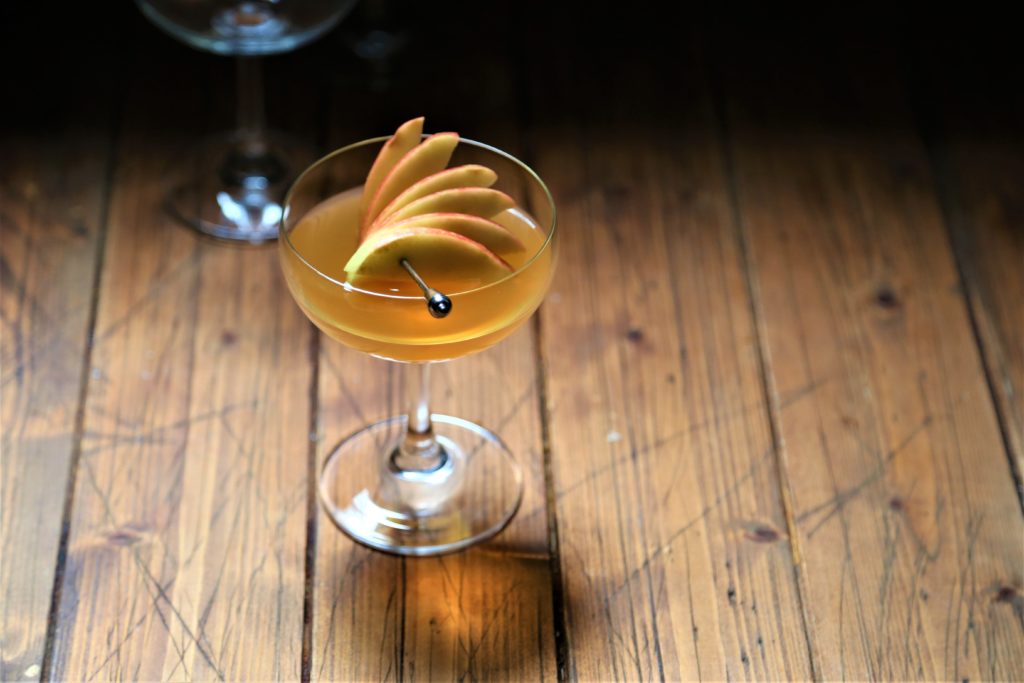 cocktail with apple fan garnish in coupe glass. Empty glass visible in background.