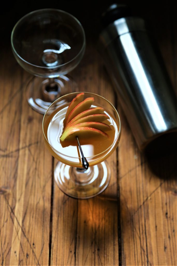 cocktail in couple glass with apple garnish. Cocktail shaker to side. Empty glass in background.
