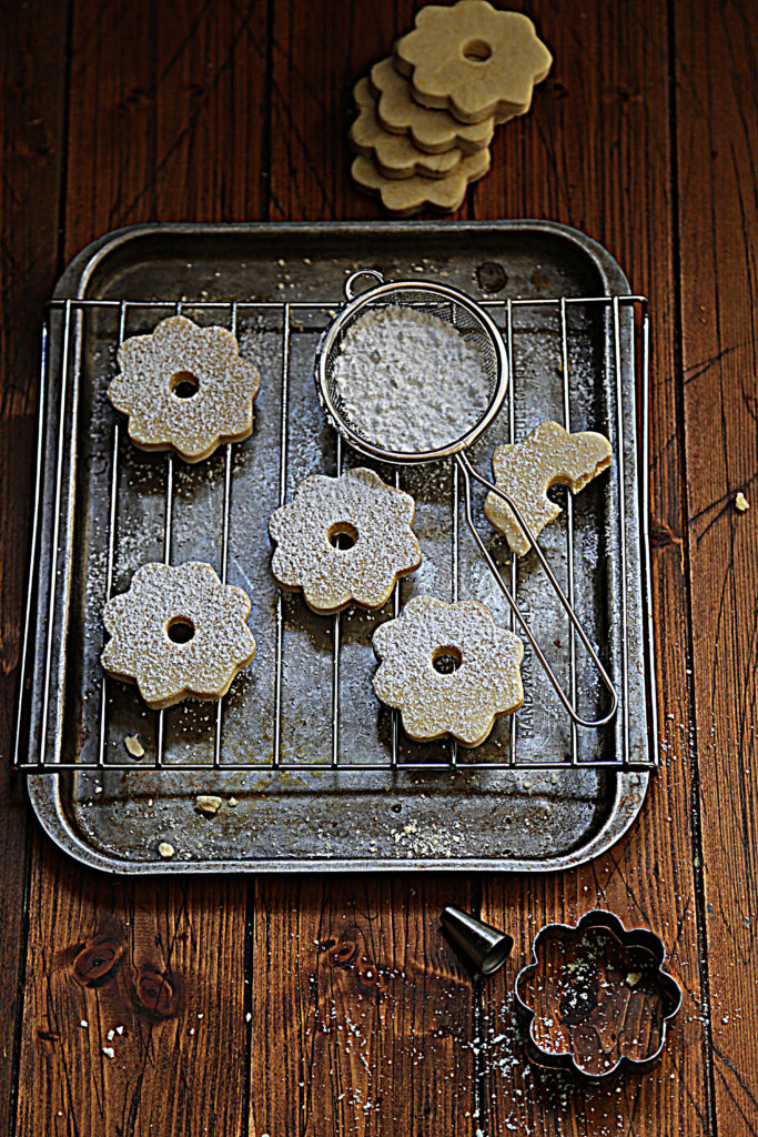 shortbread cookies on baking sheet with cooling rack. Powdered sugar in sieve on rack. Cookie cutter and decorating tip in front.