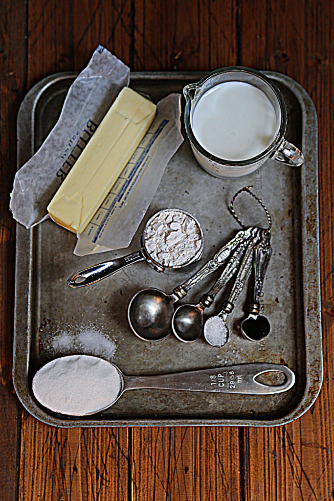 baking sheet with stick of butter, measuring spoons, measuring cup with flour, glass jar of milk, measuring spoon with sugar. 