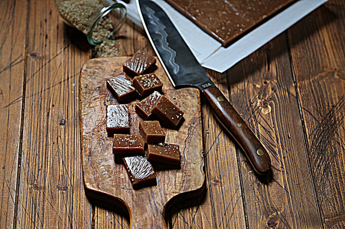 cutting board with squares of caramel scattered. Knife resting on corner. Baking pan of caramels visible in background.