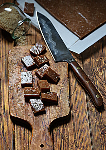 cutting board with squares of caramel scattered. Knife resting on corner. Baking pan of caramels, glass jar of salt in background.