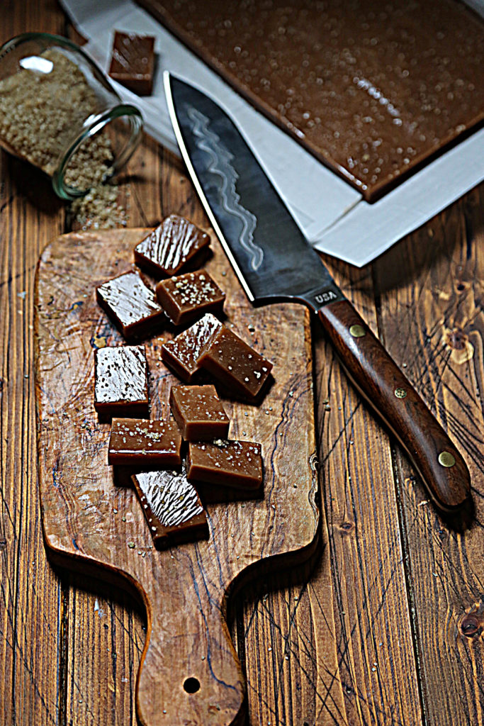 cutting board with squares of caramel scattered. Knife resting on corner. Baking pan of caramels, glass jar of salt in background.
