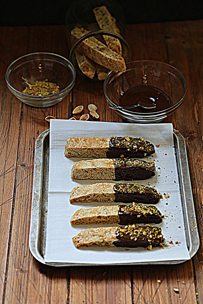 5 chocolate dipped biscotti on baking sheet lined with parchment paper. Glass jar of plain biscotti, glass bowl of chocolate, glass bowl of crushed pistachios above baking sheet.