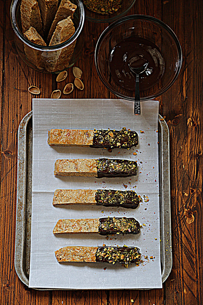 baking sheet covered with parchment paper, 5 chocolate dipped biscotti on paper. Small glass bowl of chocolate with spoon and pistachio shells above.