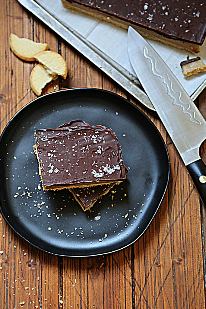black plate with 2 stacked squares of millionaire's shortbread. Shortbread cookie pieces above plate. Knife laying crosswise on baking sheet with additional shortbread. 