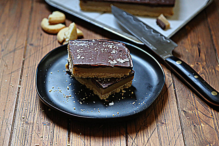 black plate with 2 squares of caramel shortbread stacked. Baking sheet with additional shortbread and knife resting on sheet above plate.