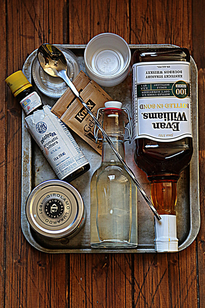 baking sheet with bottle of bourbon, bottle of simple syrup, jar of cherries, bottle of bitters, bar spoon, packet of gelatin, and plastic containers.