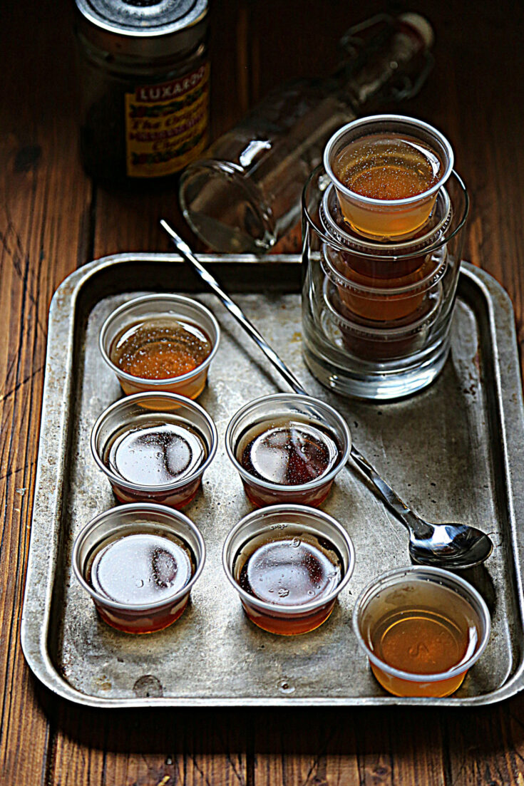 baking sheet with small plastic cups of jello shots. Bar spoon and glass with stacked cups of jello shots.