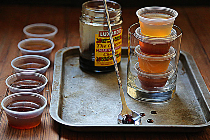 baking sheet with jar of cherries, bar spoon, glass with stacked jello shots. Jello shots to side of baking sheet. 