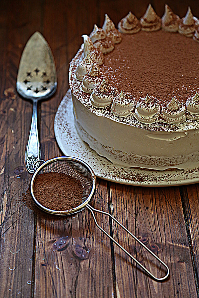 layered cake on white cake plate dusted with cocoa powder. Sieve with cocoa and cake server to side.