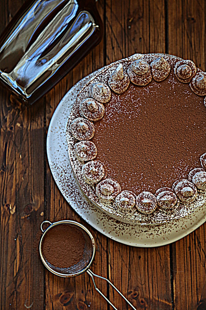 frosted cake on white white cake plate. Sieve with cocoa powder, bottle of amaretto to side. 