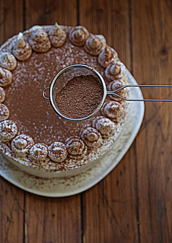 frosted cake on white cake plate. Sieve with cocoa powder being held above cake.