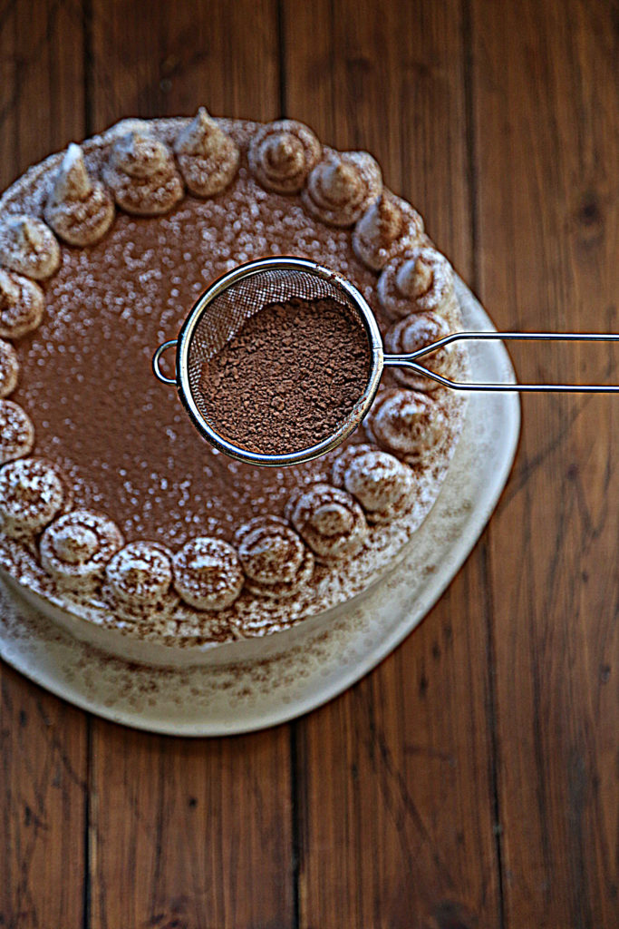 frosted cake on white cake plate. Sieve with cocoa powder being held above cake. 