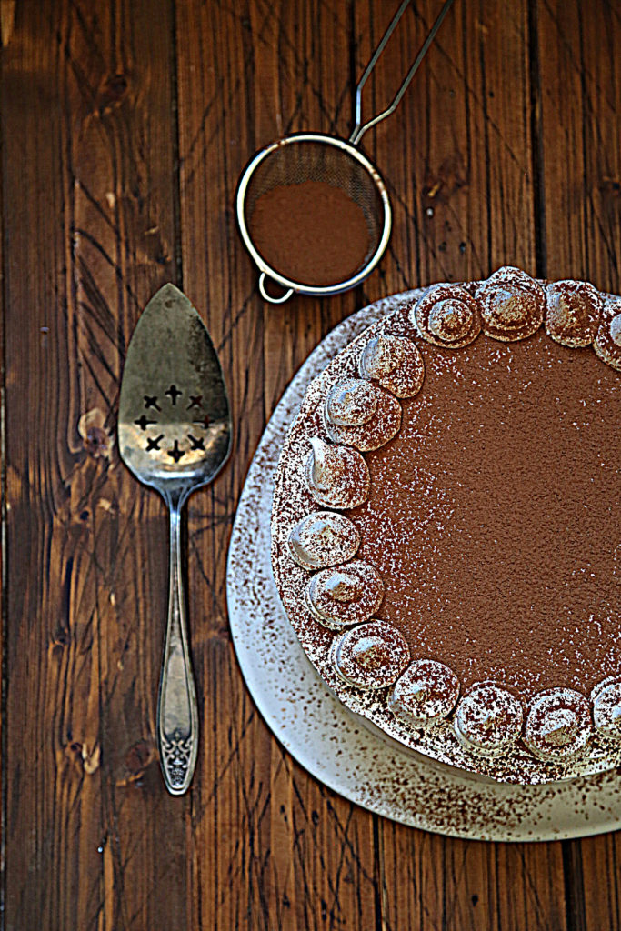 cake on white cake plate. Top dusted with cocoa powder. Cake server and sieve with cocoa powder to side. 