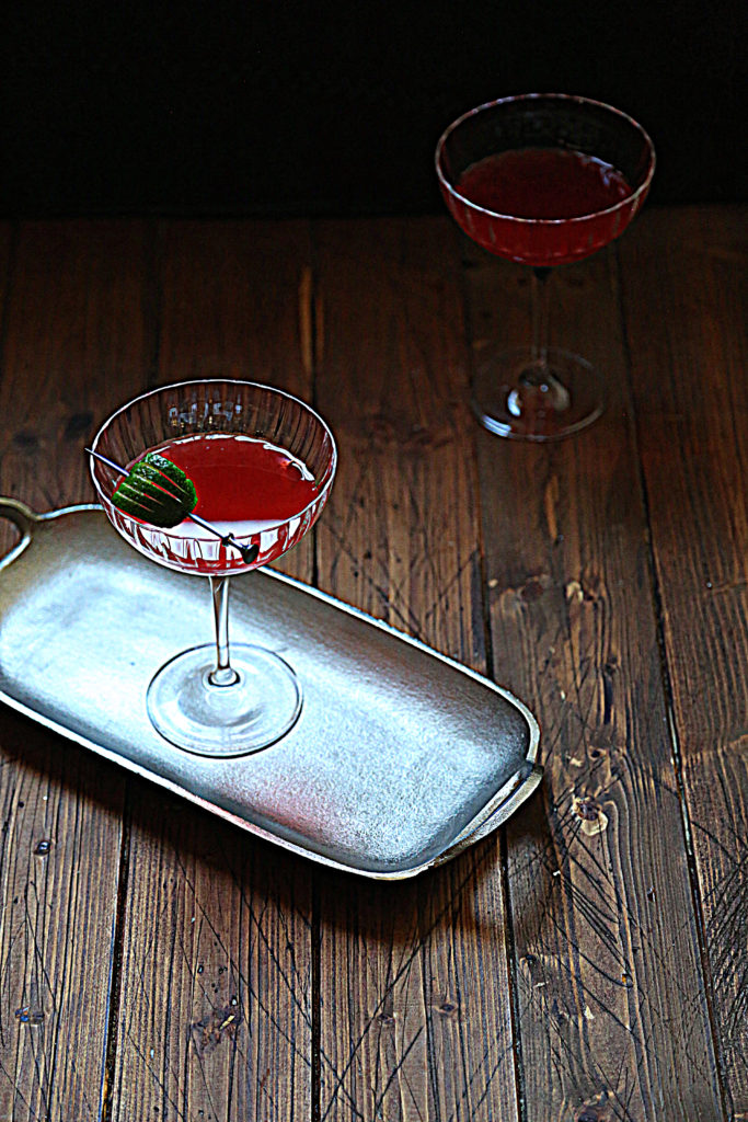 silver tray with pink cocktail in coupe glass with lime garnish, second glass visible behind tray.